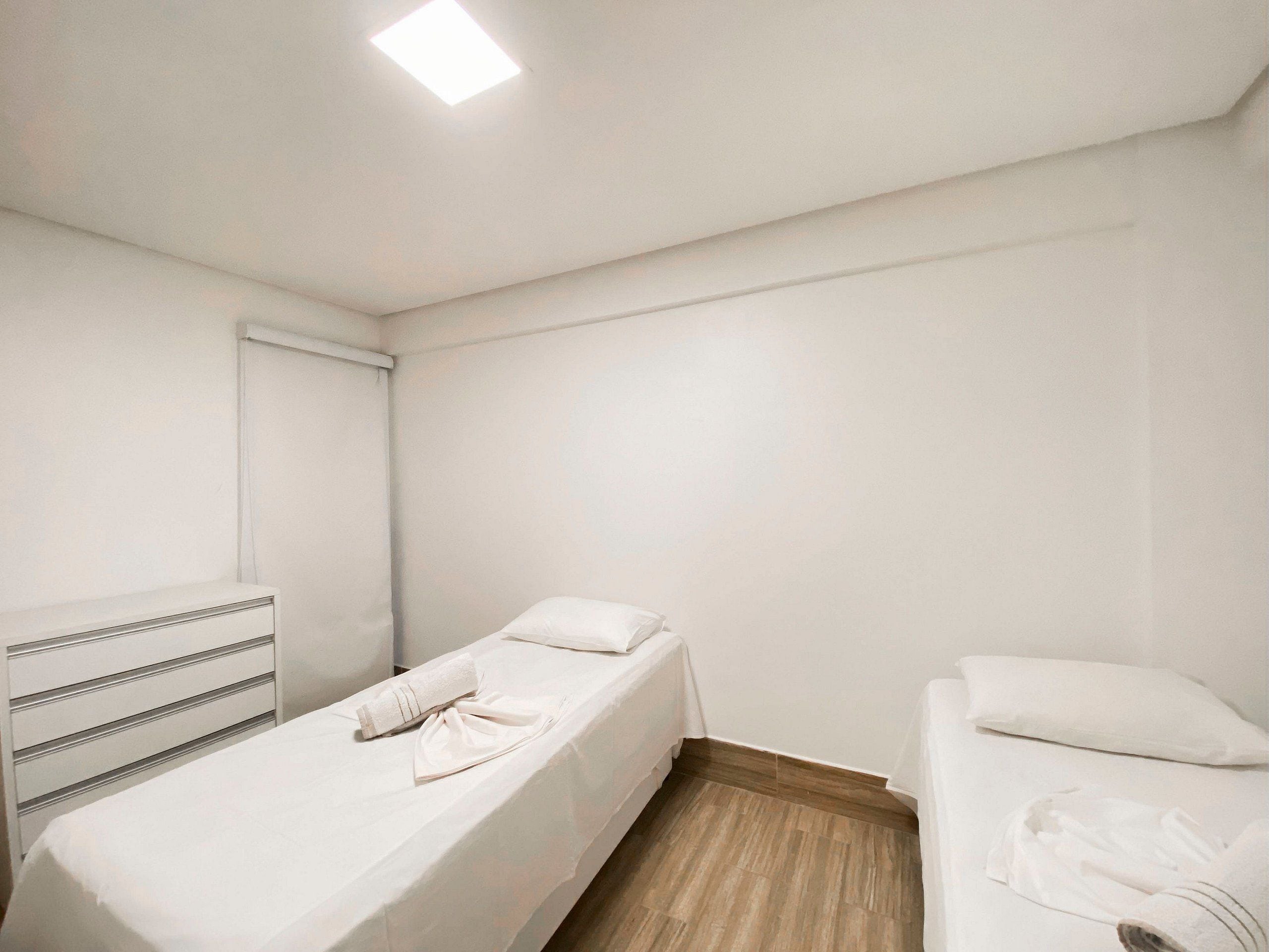 A small, clean room with two single beds, a white headboard, a wooden floor, and a ceiling light.