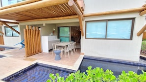Un área de piscina con bordeado de azulejos, un porche cubierto con mesa y sillas, y vista al interior a través de un puerta de vidrio.