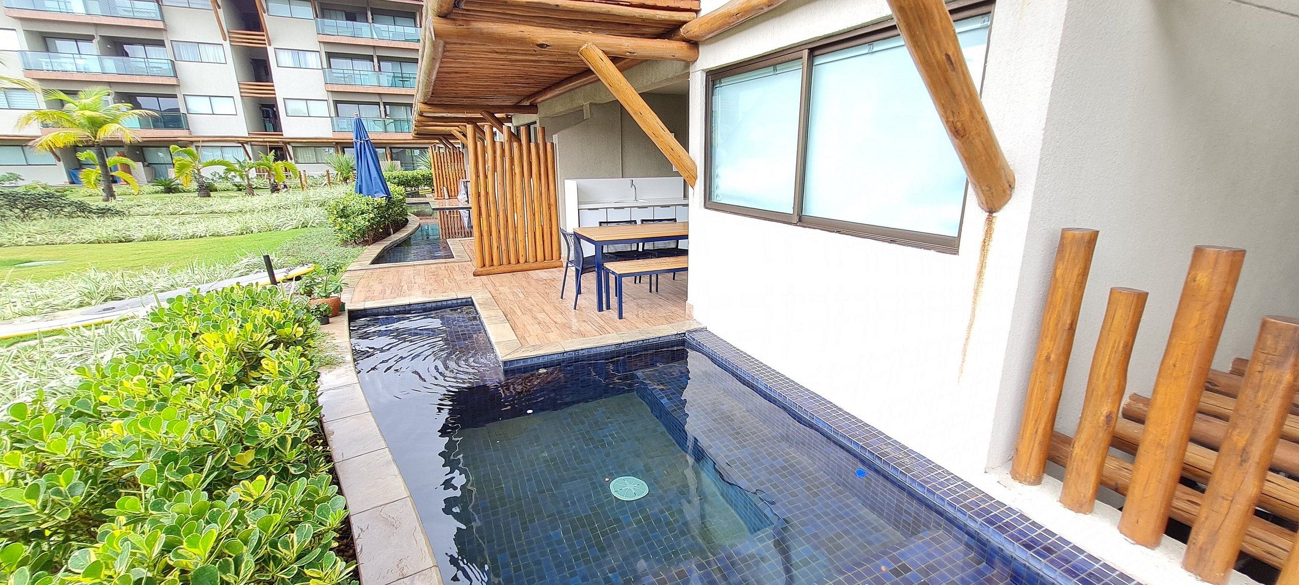 A pool area with a wooden deck, a table, and chairs, surrounded by greenery and a modern building in the background.
