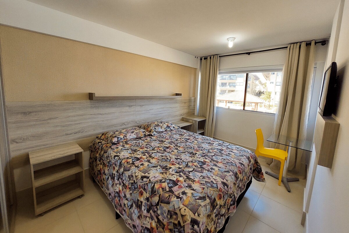 A bedroom with a bed, a bedside table, a chair, a TV, and a window with curtains.