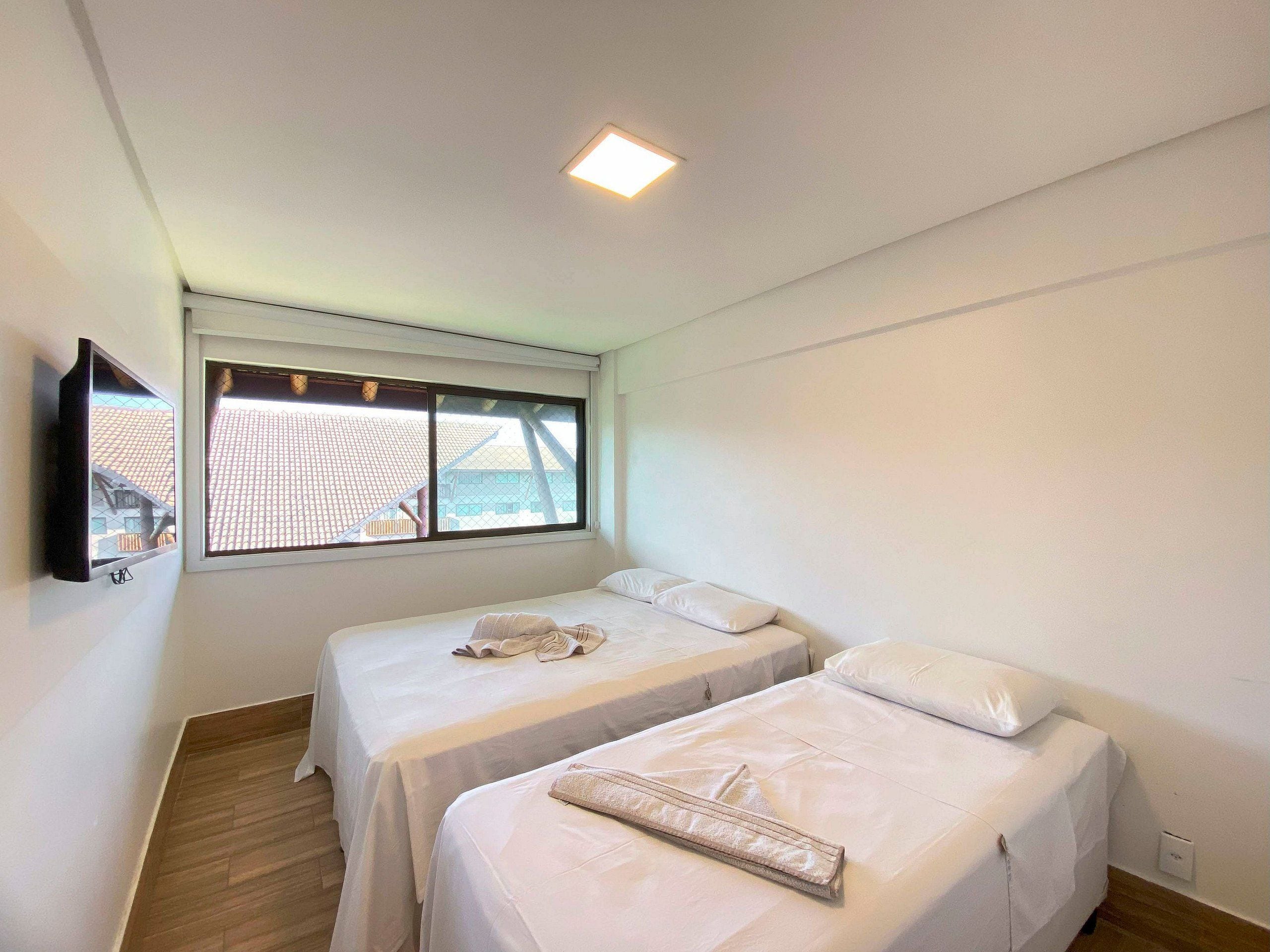 A room with two beds, a window with a view of buildings, a mounted TV, and a ceiling light.
