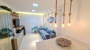 A modern living room with a sofa, a coffee table, a TV, and a dining area with a table and chairs. - Flats no La Fleur Polinésia Residence (Ipojuca)