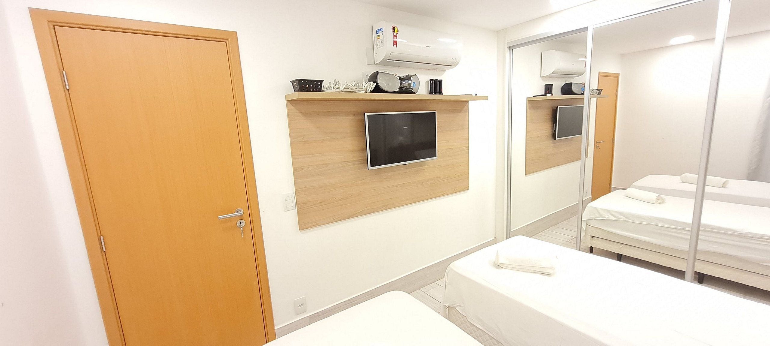 A hotel room with a wooden door, a mounted TV, a bedside table, a bed, and a mirror reflecting the room.