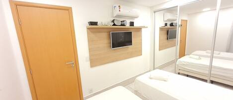 A hotel room with a wooden door, a mounted TV, a bedside table, a bed, and a mirror reflecting the room.