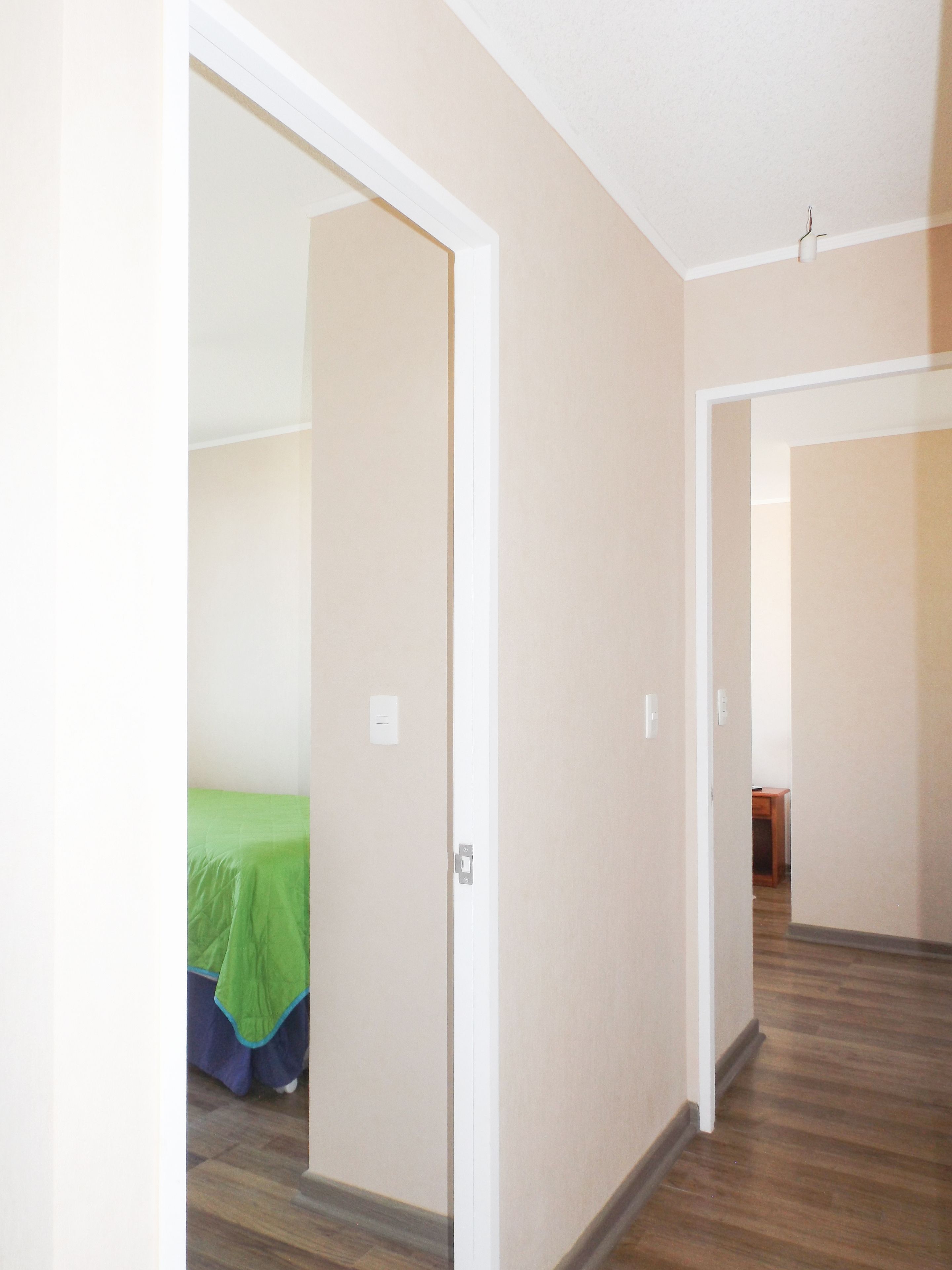 A hallway with beige walls and wooden flooring, leading to a room with a bed covered in green and blue bedding.
