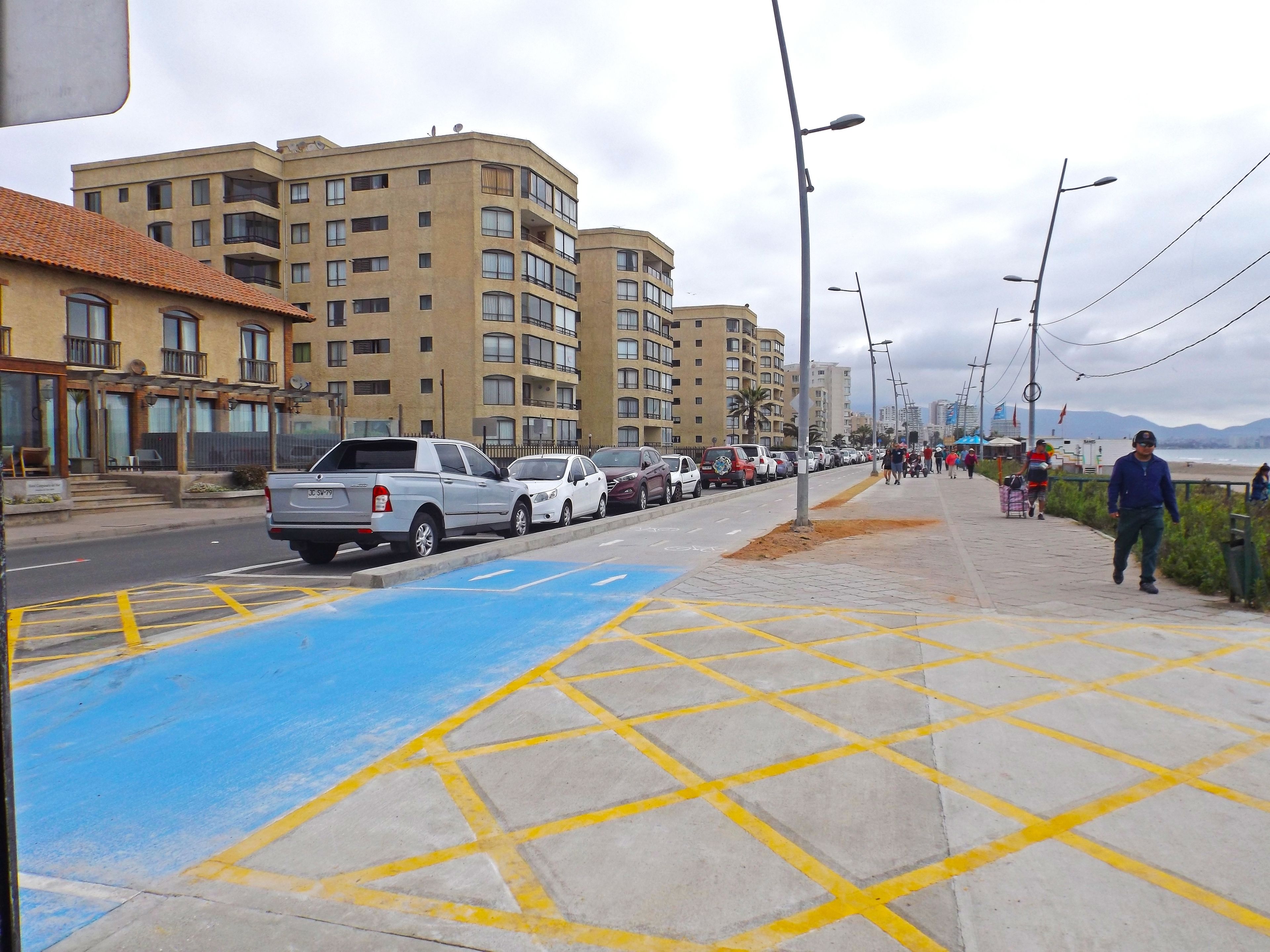 Una calle con coches estacionados, un carril bici azul y una acera con peatones.