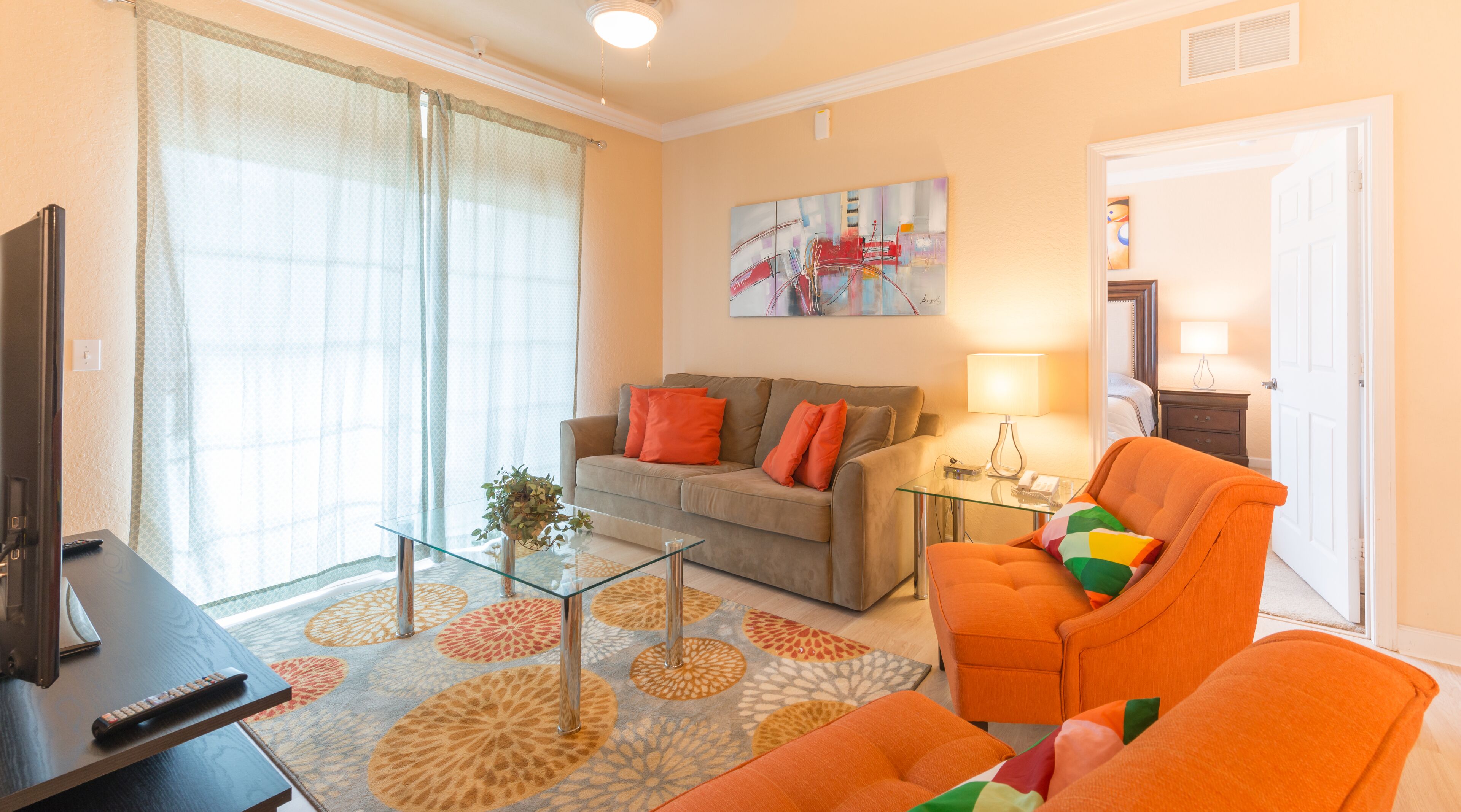 Una sala de estar con sofá, sillón y mesa de centro. Hay un televisor en un mueble bajo, una lámpara y un cuadro en la pared.