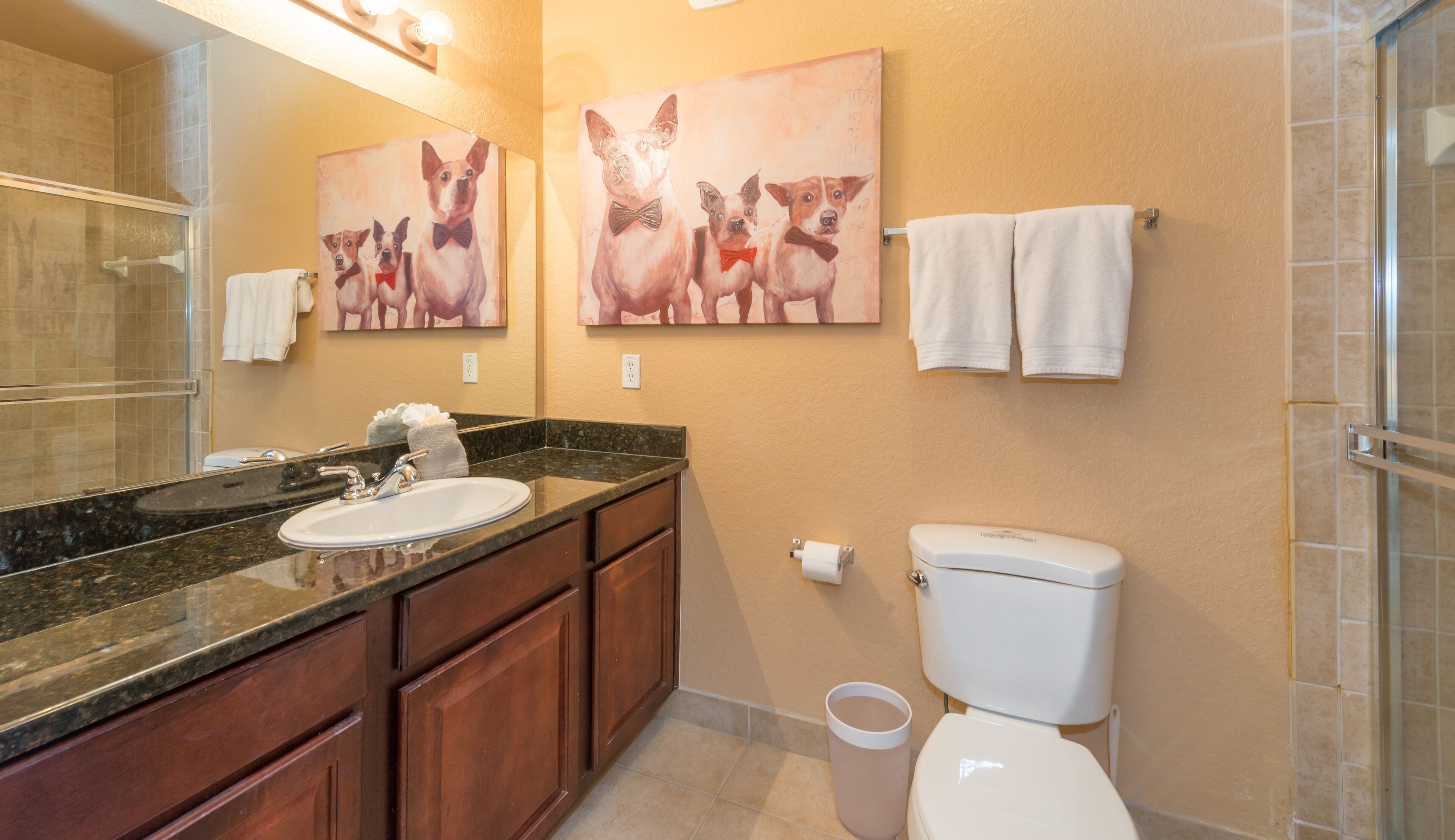 A bathroom with a sink, mirror, toilet, and shower. There are towels hanging on the wall and a painting of dogs.