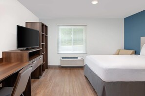 A hotel room with a bed, a desk with a television, a window with blinds, and an air conditioning unit.