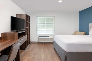 Habitación de hotel con cama, escritorio con televisor, ventana con persianas y aire acondicionado.