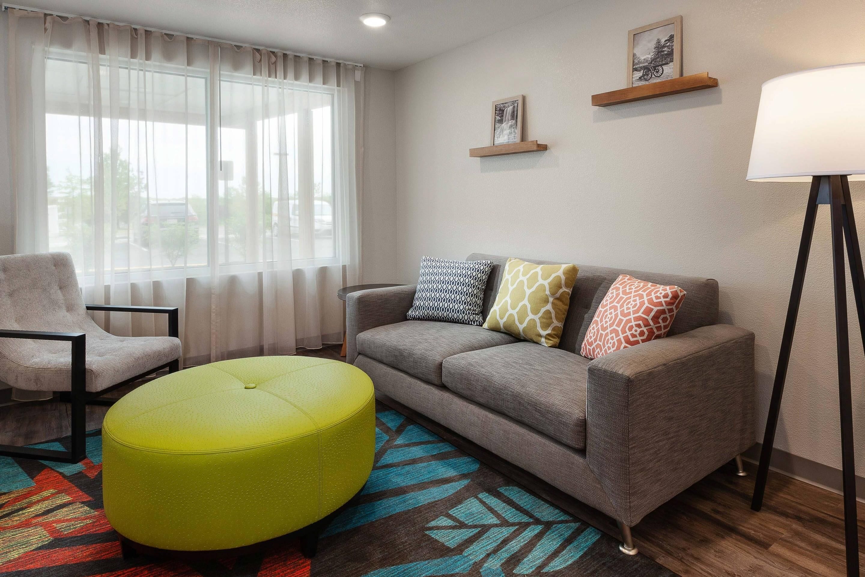 A living room with a grey sofa, a yellow ottoman, a grey armchair, a patterned rug, and a floor lamp.