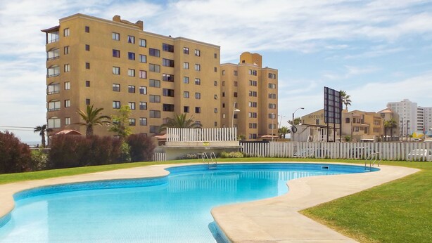 A swimming pool with a concrete deck, surrounded by a fence and greenery. In the background, there is a multi-story hotel building. - Playa Blanca (La Serena)