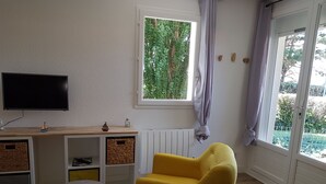 Ein Zimmer mit Holztisch, Flachbildfernseher, gelbem Sessel, Fenster mit Blick ins Grüne und Heizkörper.