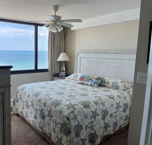 A bedroom with a large bed, a ceiling fan, a nightstand with a lamp, and a view of the ocean through a window.