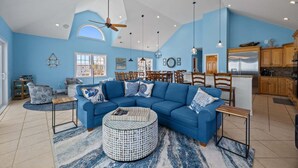 A spacious living room with a blue sofa, a round coffee table, and a dining area with wooden chairs. - 5849- Mari-Time, Oceanfront 8BR Luxury: Theater, Pool & Elevator (South Nags Head)