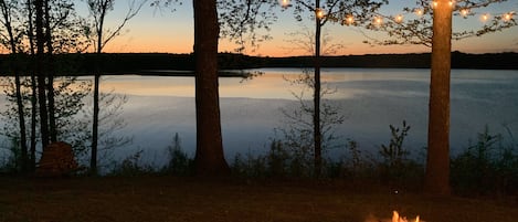 A lakeside view with trees, a fire pit, and string lights.