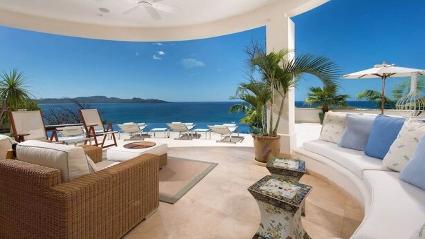 Uma área de estar ao ar livre e ensolarada com um sofá branco curvo, mobiliário de vime e vista para o oceano.