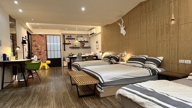 A modern hotel room with a wooden floor, two beds with striped bedding, a desk with a chair, a small table, and a green chair.