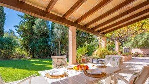 An outdoor patio with a wooden pergola, white tables, and chairs, surrounded by lush greenery and trees.