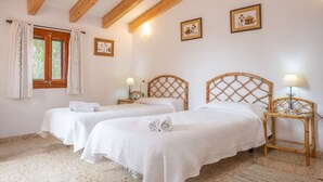 A room with two beds, wooden beams on the ceiling, a window with curtains, and wall-mounted lamps.