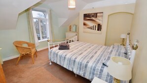 A bedroom with a bed, a chair, a bedside table with a lamp, a window with curtains, and a painting on the wall. - Flintrock House -  a house that sleeps 9 guests  in 5 bedrooms (Sheringham)