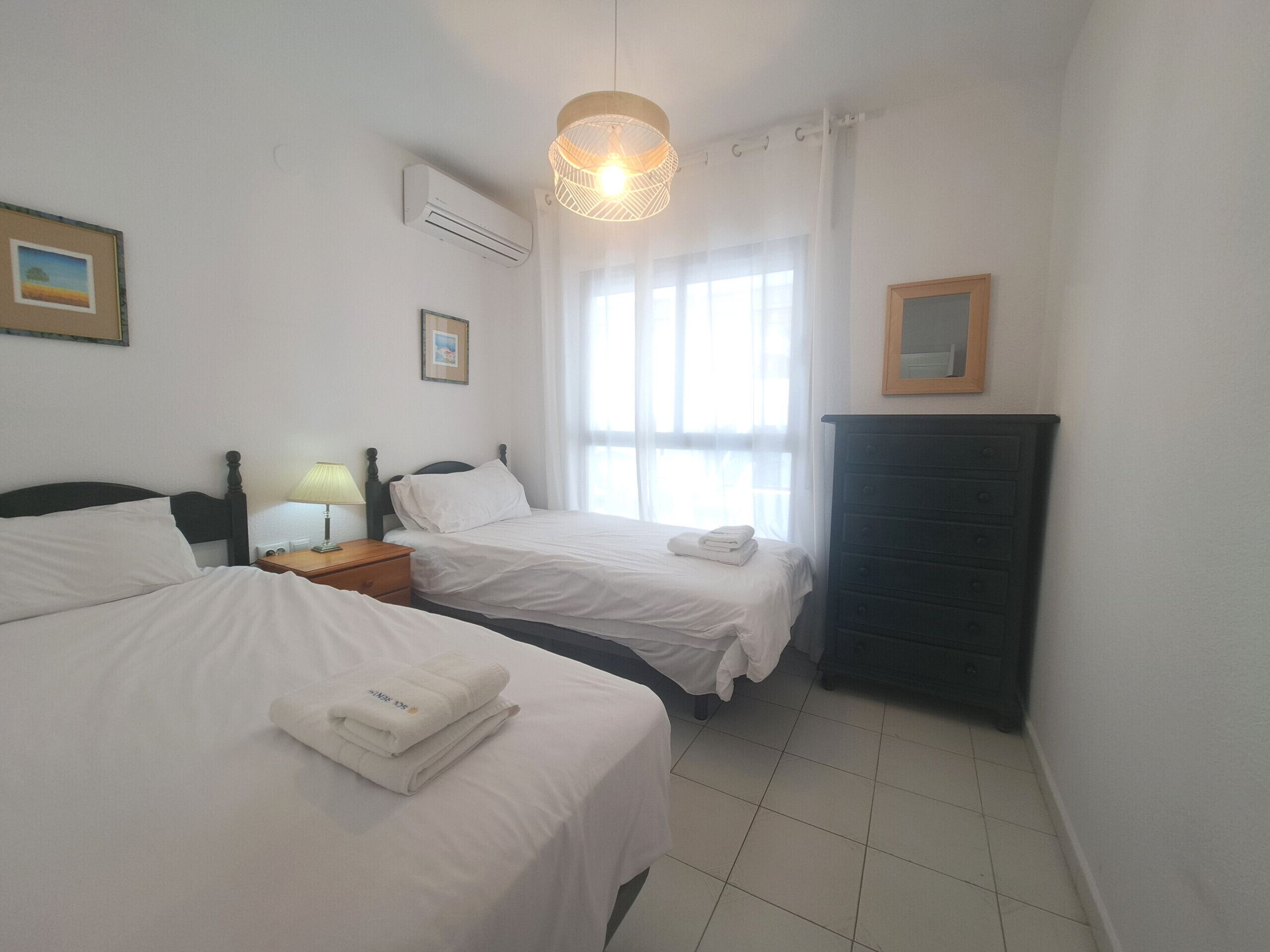 A hotel room with two beds, a nightstand with a lamp, an air conditioning unit, a dresser, and a window with curtains.