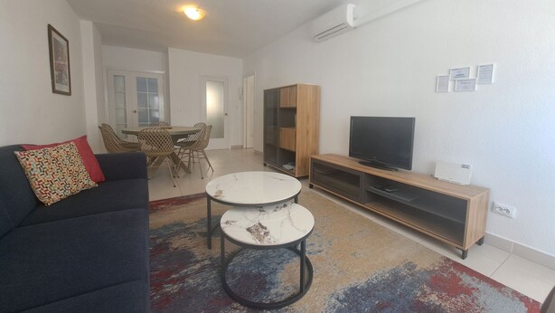 Un salon moderne avec un canapé foncé, deux tables rondes, un téléviseur posé sur un meuble en bois et un coin repas comprenant des chaises et une table.
