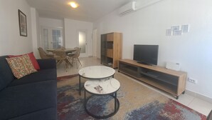 A modern living room with a dark sofa, two round tables, a TV on a wooden stand, and a dining area with chairs and a table.
