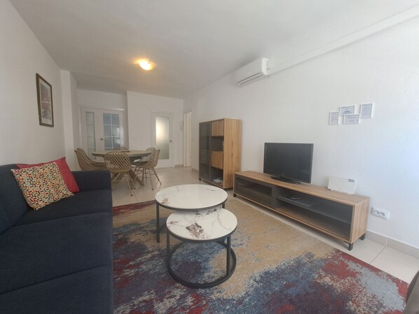 A modern living room with a dark sofa, two round tables, a TV on a wooden stand, and a dining area with chairs and a table. - Ref: 299 Beachfront 4 bedroom apartment with pool in Fuengirola (Fuengirola)