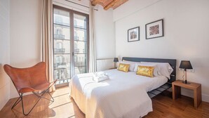 Quarto com cama, cadeira, mesa de cabeceira e janela com vista para prédios.