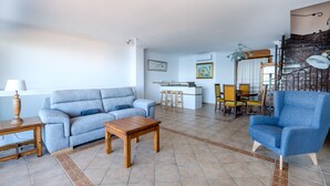Una sala de estar con sofá, mesa de centro y un sillón azul. Se observa una zona de cocina con mostrador y taburetes. Se ve una escalera con barandilla.