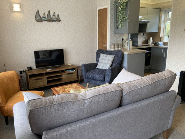 A living room with a sofa, armchair, TV, wooden entertainment unit, and a kitchen area with cabinets and a countertop.