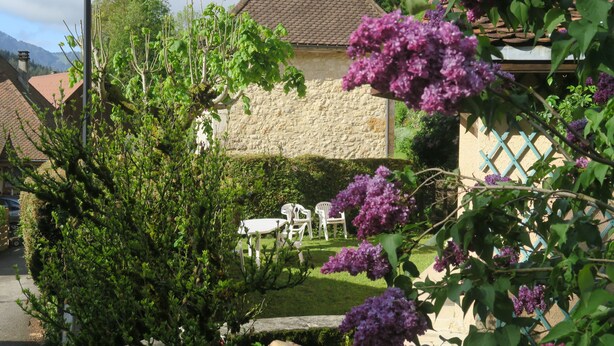 Una casa de piedra con tejado de tejas, un jardín con sillas blancas y flores púrpuras.