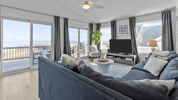 A spacious living room with a large sofa, a television, and a balcony with a view of the beach. - 1661 - The Best Nest - Oceanfront Corolla | Pool, Putt-Putt & Hot Tub (Corolla)