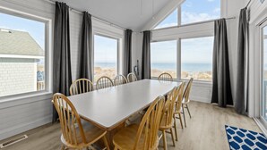 A room with a large table and chairs, large windows with curtains, and a view of the beach. - 1661 - The Best Nest - Oceanfront Corolla | Pool, Putt-Putt & Hot Tub (Corolla)