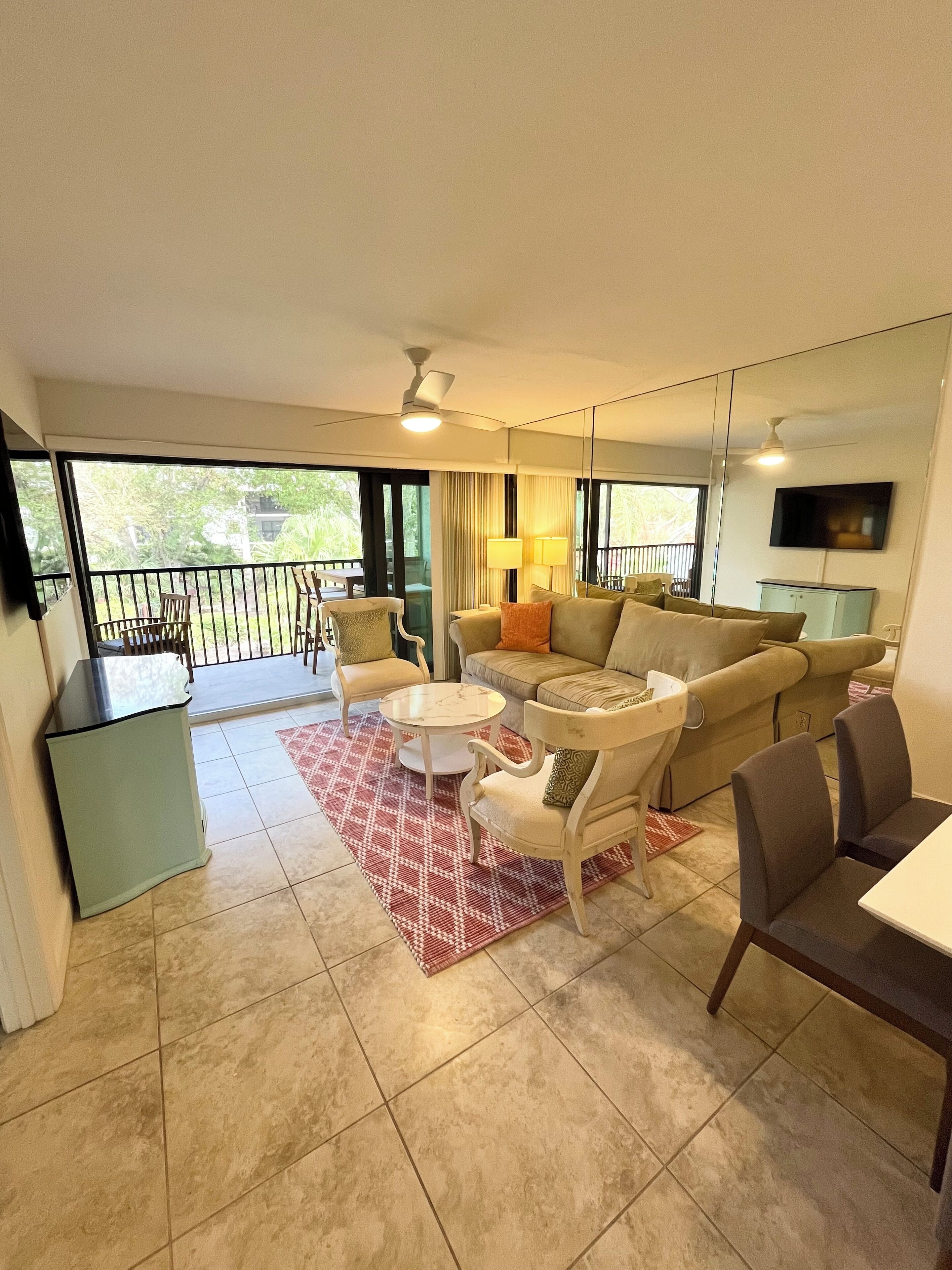 A spacious living area with a sofa, chairs, a coffee table, and a TV. There is a balcony with a railing and a view of greenery.