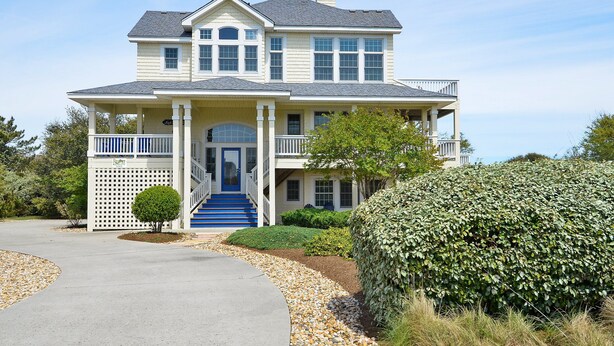 A two-story house with a front porch, a covered entryway, and a staircase leading to the main entrance. - 2304 - High Spirits - Pine Island Retreat | Pool, Hot Tub, Near Beach (Corolla)