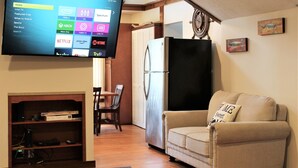 Un soggiorno con una TV a schermo piatto che mostra l'interfaccia di un servizio di streaming, un frigorifero, un divano, un tavolo da pranzo con sedie e una libreria.
