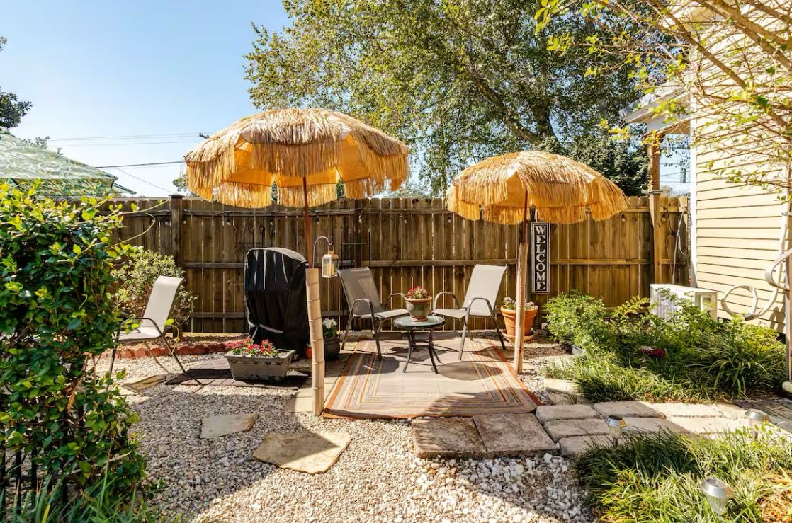 Un patio trasero con valla de madera, dos sombrillas de paja, una parrilla y mobiliario de exterior.