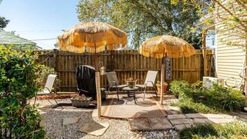Un patio trasero con valla de madera, dos sombrillas de paja, una parrilla y mobiliario de exterior.