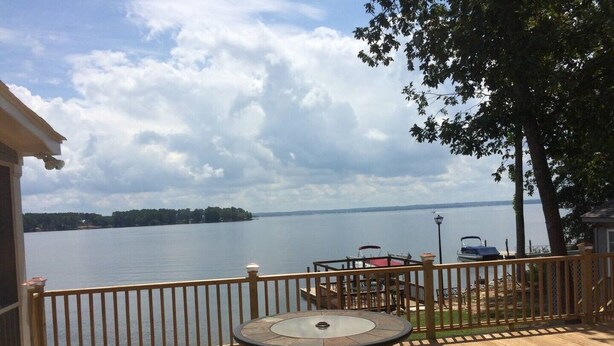 A wooden deck with a round table and chairs, overlooking a calm lake with boats and a distant shoreline.