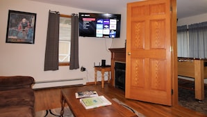 A living room with a flat-screen TV mounted on the wall, a brown couch, a wooden coffee table with books, a fireplace, and a wooden door. - Cooperstown Baseball Rentals - Lakeview Pines (Maryland)