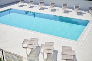 A rectangular swimming pool with clear blue water, surrounded by white tiled flooring and beige sun loungers. - West Village Appartamento - NUOVO con piscina (Jesolo)