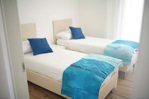 Two single beds with white bedding and blue pillows in a room with wooden flooring. - West Village Appartamento - NUOVO con piscina (Jesolo)