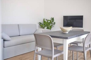 A modern living room with a grey sofa, a dining table with four chairs, and a flat-screen TV mounted on the wall. - West Village Appartamento - NUOVO con piscina (Jesolo)