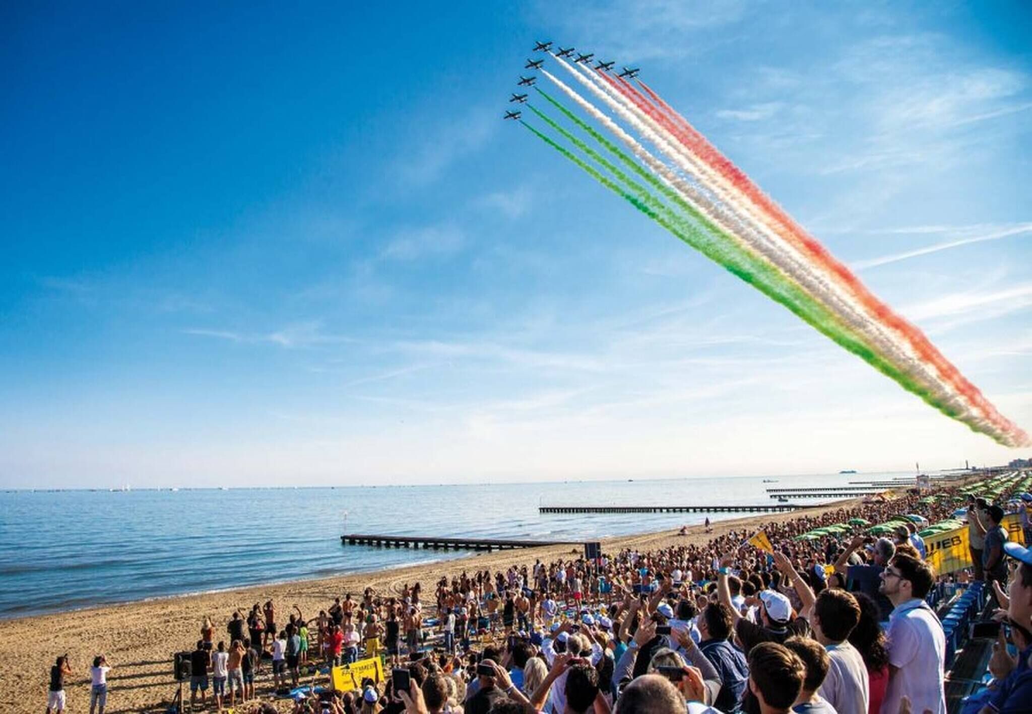 Une plage où une foule observe une formation d’avions qui tracent des traînées de fumée colorée dans le ciel.