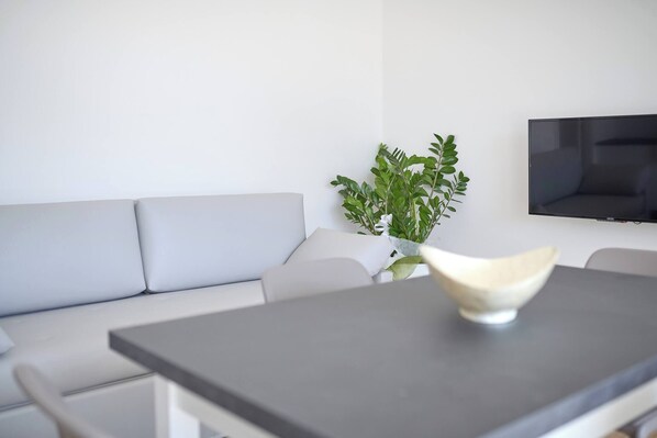 A modern living room with a grey sofa, a dining table with a white bowl, a potted plant, and a flat-screen TV mounted on the wall. - West Village Appartamento - NUOVO con piscina (Jesolo)