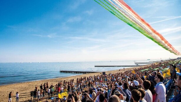 A beach with a crowd watching a formation of aircraft creating colored smoke trails.