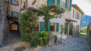 A cobblestone street with a building featuring a balcony with green shutters, a small table with chairs, and a view of a mountain in the background.
