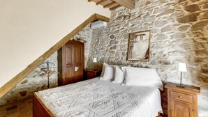 Quarto com teto inclinado, paredes de pedra, cama com roupa de cama estampada, móveis de madeira e um quadro na parede.